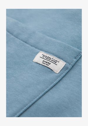 Lyst blå bomuldsstof med en tekstureret overflade, der har et hvidt mærke prægede med "DIN ENESTE GRÆNSE ER DIN HJERNE" og "OMBRE Siden 1985."