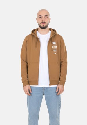 Hombre vestido con una sudadera marrón con cremallera y estampado gráfico blanco, camiseta blanca y vaqueros azul claro, posando frente a un fondo liso.