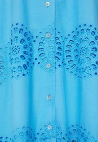 Chemise bleu clair ornée d'une broderie florale en bas, détaillée avec des motifs circulaires, et des boutons décoratifs sur le devant.