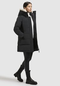 Sort pufferjakke med aftagelig faux fur-forsynet hætte, lynlås-lukning og to sidelommer; parret med sorte leggings og ankelstøvler.