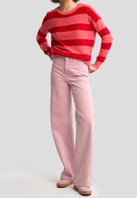 Červený a růžový pruhovaný pletený svetr s dlouhými rukávy, doplněný o vysoké světle růžové džíny se širokými nohavicemi a béžovou obuv.