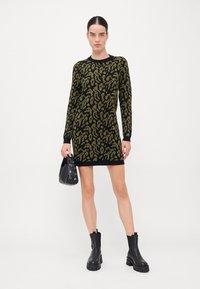 Strikket kjole med grønn leopardmønster på svart bakgrunn, lange ermer, ribbede mansjetter og en tettsittende silhuett. Tilbehøret inkluderer en svart håndveske.