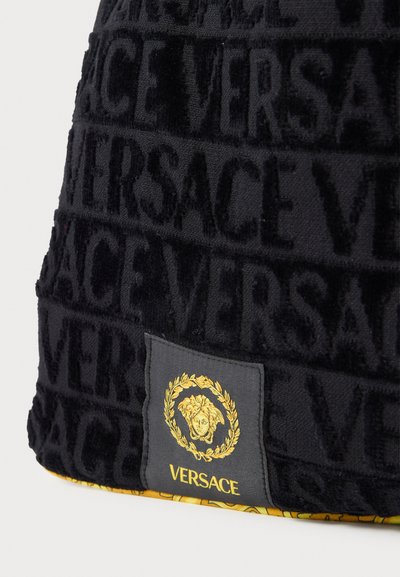 Crni teksturirani materijal s reljefnim uzorkom "VERSACE", s kontrastnom etiketom na kojoj se nalazi zlatni grb i detalji logotipa.
