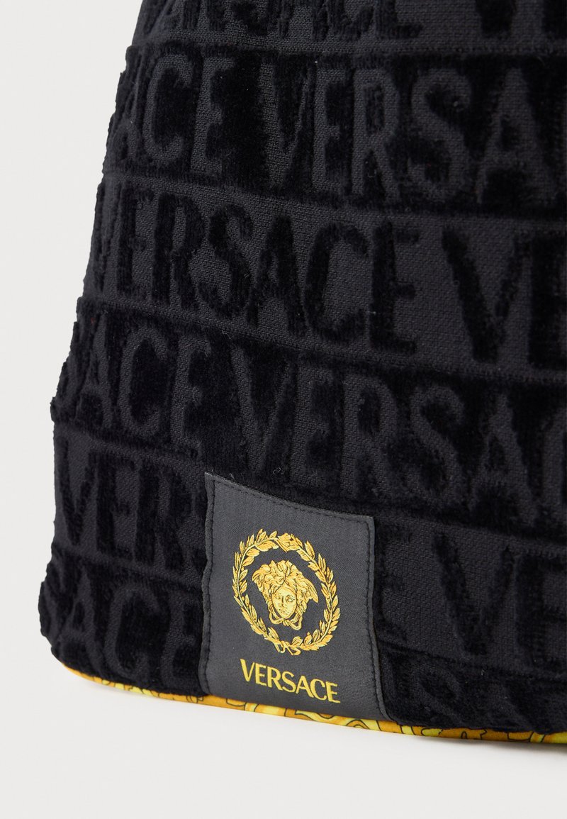 Sort tekstureret stof med et præget "VERSACE" mønster og et kontrasterende mærke med en guldfarvet emblem- og logodetalje.