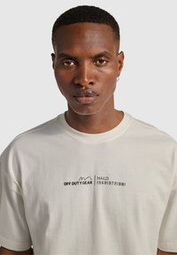 Jasno-szara koszulka z krótkim rękawem z nadrukiem logo w kolorze czarnym, zawierająca napisy "OFF DUTY GEAR" oraz "HALO 196819721981". Miękka bawełniana tekstura.