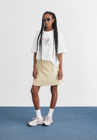 Vit grafisk t-shirt med sneakertryck, beige denimkjol och rosa träningsskor. Flätat hår och mörka solglasögon fulländar looken.