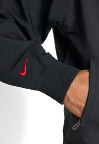 Veste noire en matière légère et texturée. Comprend des poignets élastiques et une poche zippée. Logo Nike rouge sur le poignet.