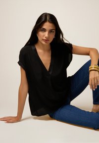 Blusa nera con scollo a V e maniche corte a palloncino, realizzata in tessuto leggero. Indossata con jeans blu, caratterizzati da una superficie liscia.