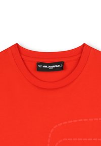 T-shirt en coton rouge avec col rond, présentant une texture douce et un subtil motif de contour cousu sur le devant. Étiquette sur le col.