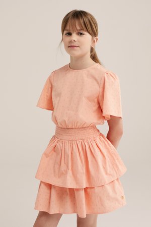 Mlada djevojka stoji s rukama iza leđa, nosi breskvaste haljine kratkih rukava s naboranim strukom i slojevitom suknjom na jednobojnoj pozadini.