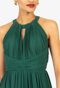 Abito verde con tessuto plissettato, design con collo halter e vita aderente. La texture è liscia e fluida, accentuando il drappeggio.