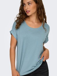 Ung kvinne med krøllete hår iført en lyseblå kortermet skjorte, holder i skjortekanten og ser litt mot venstre.