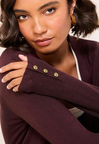 Cardigan bordeaux avec une texture côtelée, des manches longues et trois boutons en forme de crâne dorés sur les poignets.