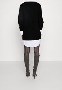 Maglione nero oversize con bordo a coste, indossato sopra una camicia bianca. La modella indossa collant neri e stivali con tacco grigio fino al ginocchio.