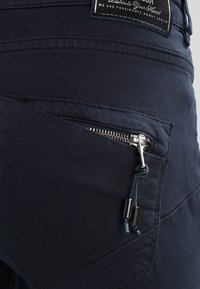 Tmavě modré látkové kalhoty s kapsou na zip, která má stříbrný zip a modrý šňůrkový akcent. Materiál má texturovaný povrch.