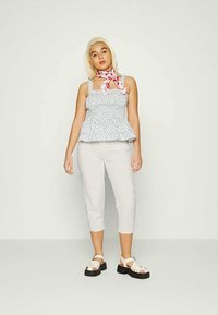 Mulher com cabelo loiro, vestindo um top sem mangas branco com flores, calças brancas cropped, sandálias de plataforma brancas e um lenço de pescoço com padrão rosa.