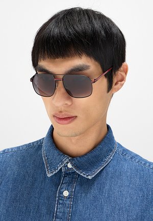 Giovane uomo con capelli corti e neri che indossa occhiali da sole rettangolari scuri e una camicia di jeans blu con bottoni.