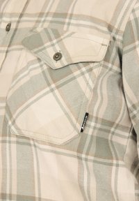 Close-up van borstzak van een beige en groene geruite blouse met knoopflap en klein merklabel op de stof.