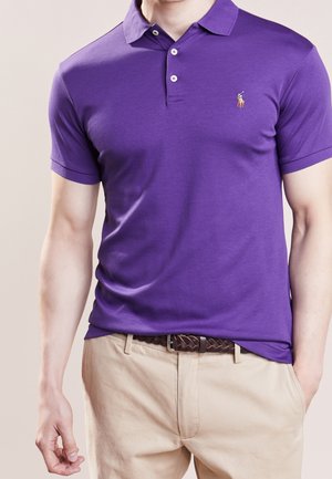 Polo shirt viola a maniche corte con colletto a coste, patta con due bottoni e un logo ricamato discreto. Indossato con pantaloni beige chiaro.