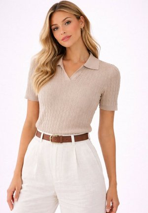 Femme aux longs cheveux blonds portant un polo tricoté beige à manches courtes et un pantalon blanc taille haute avec une ceinture marron, debout devant un fond blanc.