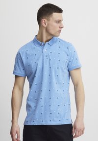 Lyseblå poloshirt laget av bomull, med et spredt mønster av mørkeblå prikker og diamanter, med krage og korte ermer.