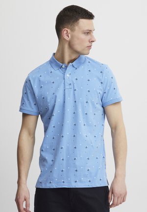 Camisa polo de color azul claro, hecha de algodón, con un patrón disperso de puntos y diamantes azul oscuro, con cuello y mangas cortas.