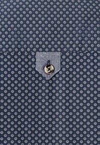 Dunkelblauer Stoff mit einem weißen Diamantmuster, einer strukturierten aufgesetzten Tasche und einem Knopfdetail in der Ecke.