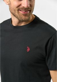 Juodas medvilninis marškinėliai su apvaliu kaklu ir mažuoju raudonu siuvinėjimu ant kairiojo krūtinės kampo. Trumpomis rankovėmis su lygiu paviršiumi.