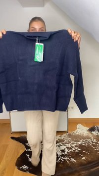 Személy, aki egy sötétkék bordázott pulóvert tart a kezében zöld címkével, egy barna és fehér tehénbőr szőnyegen áll egy világos szobában.