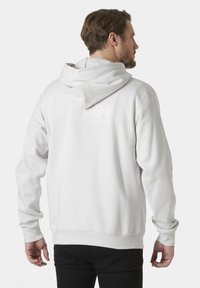 Sudadera blanca con una textura suave, presenta una gran capucha, mangas con puños y un logo sutil en la parte trasera. Combinada con pantalones negros.