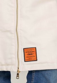 Lekka kurtka bomber w kolorze off-white z złotym zamkiem, z pomarańczową i czarną metką z informacjami o marce i specyfikacji.