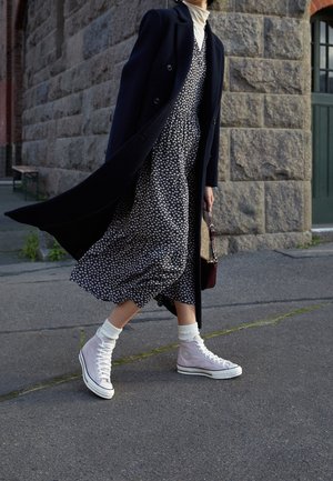 Cappotto doppiopetto nero sopra un vestito floreale scuro, abbinato a sneaker alta lilla e calzini bianchi. Borsa bordeaux in mano.