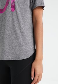 Szara, krótkosleeve koszulka sportowa z różowym wzorem graficznym, miękka struktura materiału i zaokrąglony dół. Czarne legginsy są częściowo widoczne.