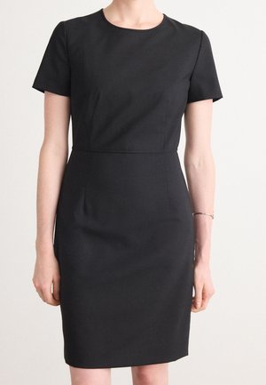 Femme portant une robe noire ajustée à manches courtes, longueur genoux, avec un col rond et un fin bracelet au poignet droit.