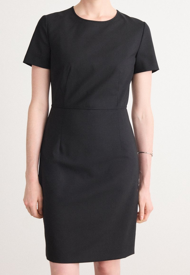 Femme portant une robe noire ajustée à manches courtes, longueur genoux, avec un col rond et un fin bracelet au poignet droit.