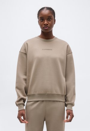 Kobieta w beżowej bluzie z długim rękawem i dopasowanych spodniach, stojąca na białym, jednolitym tle.