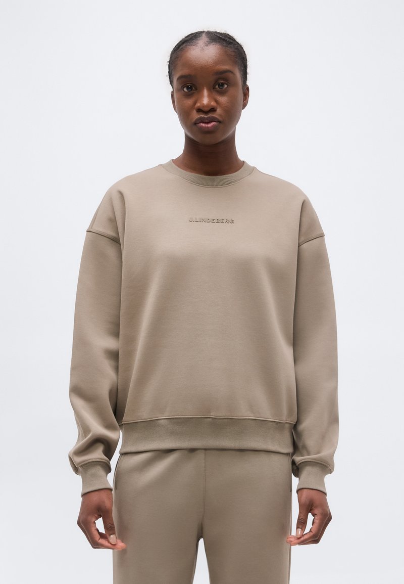 Γυναίκα που φοράει μπεζ φούτερ με μακριά μανίκια και matching παντελόνι, στέκεται μπροστά σε ένα απλό λευκό φόντο.