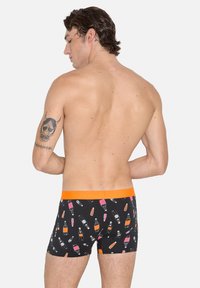 Hombre sin camiseta con un tatuaje en el brazo izquierdo, llevando bóxer negros con un patrón de botellas de licor en color naranja y rosa, cinturilla naranja, fondo blanco.