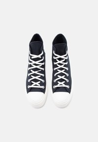 Ζευγάρι μαύρα ψηλά πάνινα παπούτσια με λευκές λαστιχένιες μύτες και λευκά κορδόνια, σε άποψη από πάνω, σε ανοιχτό γκρι φόντο.