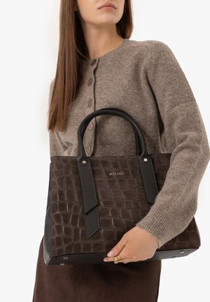 Woman in a brown buttoned sweater holding a dark brown textured handbag with black handles and a gold ring on her finger.