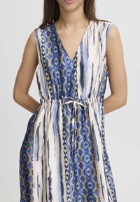 Robe sans manches avec un décolleté en V dans des motifs bleus, crème et verts. Tissu léger, taille cintrée avec un cordon de serrage, dotée de poches latérales.