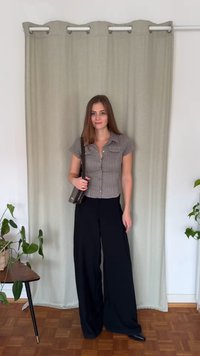 Svarte vid bukser kombinert med en rutete bluse med korte ermer. En brun håndveske henger over skulderen. Bakgrunnen har et grønt gardin.