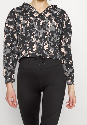 Nainen pukeutuneena mustaan, harmaaseen ja vaaleanpunaiseen camokuvioituun lyhytpukuiseen huppariin ja mustiin korkeavyötäröisiin leggingsseihin.