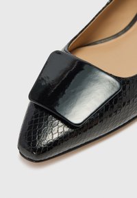 Chaussure noire à bout pointu faite en cuir verni texturé avec un accent carré noir brillant sur le dessus. Doublure intérieure lisse.