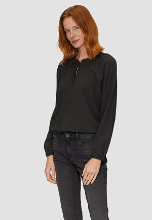 Blouse noire texturée avec un décolleté en trou de serrure et un accent noué. Manches longues avec des poignets élastiques, associée à un jean foncé.