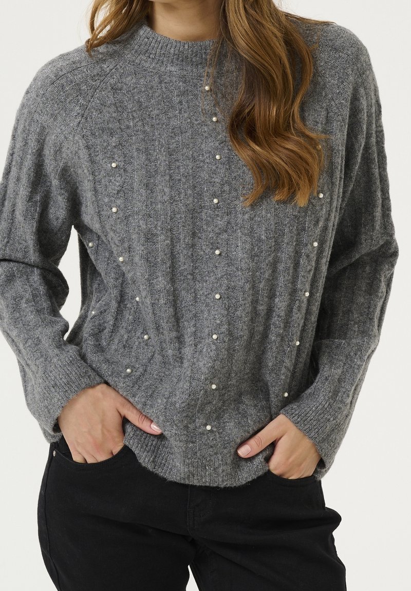Femme portant un pull gris torsadé avec de petites perles, associé à un pantalon noir, mains dans les poches, cheveux longs et ondulés.