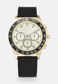 Montre chronographe dorée avec une lunette noire, un cadran crème, trois sous-cadrans et un bracelet en silicone noir. Présente des repères de minutes et des poussoirs.
