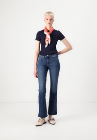 T-shirt bleu marine à manches courtes, jeans bleus évasés, et écharpe rouge à motifs nouée au cou. Chaussures beiges avec un détail de boucle.
