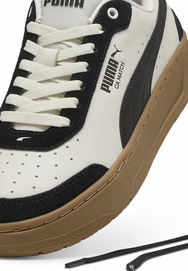 Baskets en cuir blanc avec des accents en suède noir, bout rond, semelle en caoutchouc texturé couleur tan, design à lacets et logo Puma sur le côté.