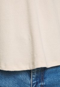 Ourlet en tissu beige reposant sur un jean en denim bleu avec des coutures visibles au niveau de la couture et de la ceinture.